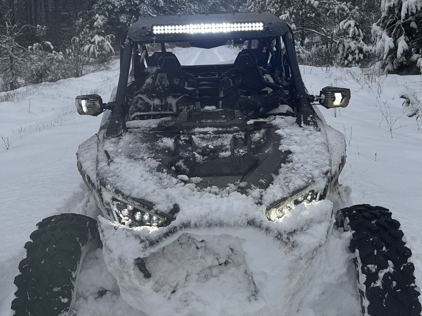 A side-by-side with its light bar on, buried nose-deep in snow on a wooded trail